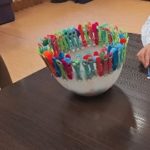 Terapia ręki - konsultacje - ćwiczenie manualne z wykorzystaniem kolorowych klamerek i pomponów umieszczonych wokół miski. Obok dziecko wykonuje zadanie na kartce.